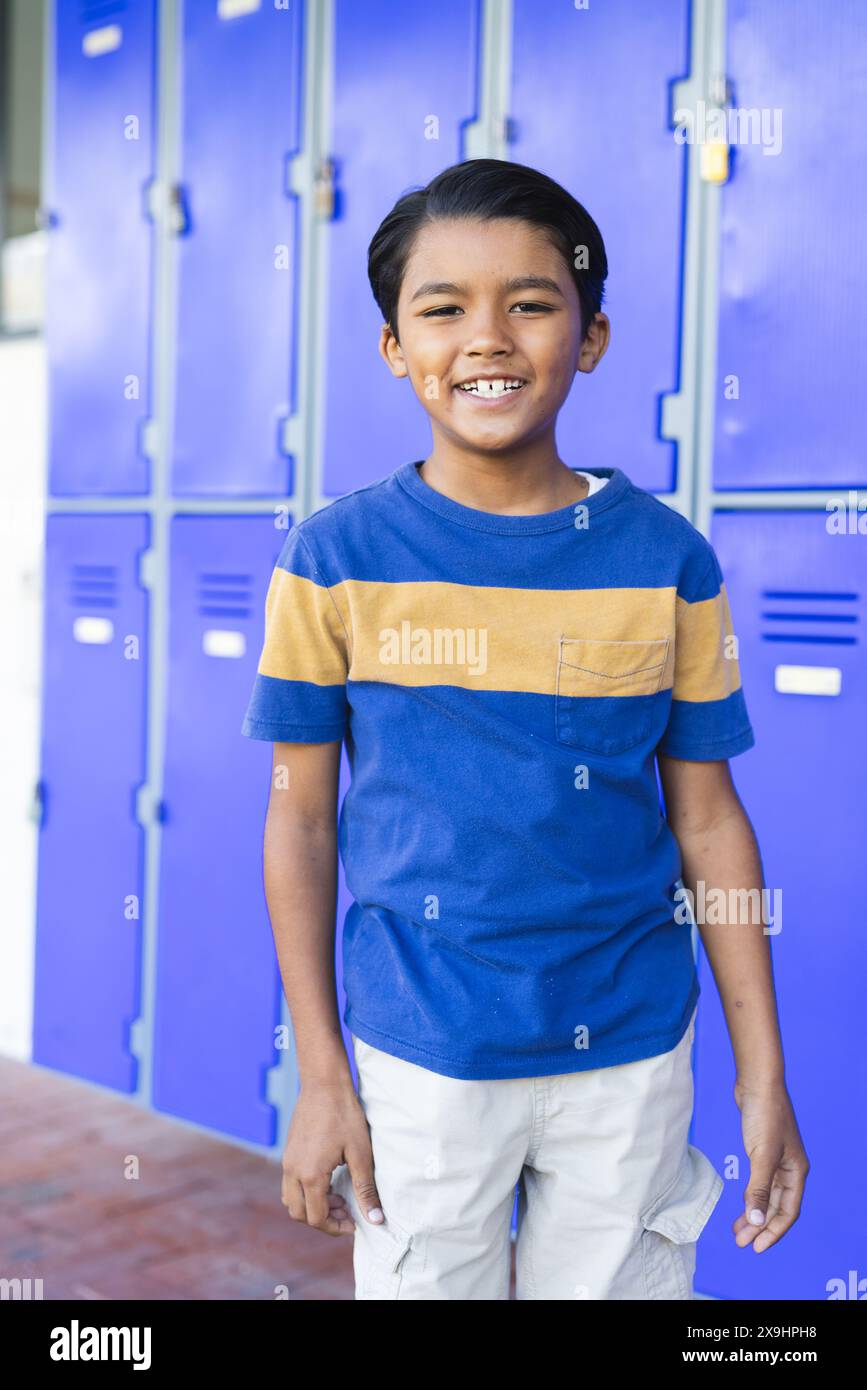 Un ragazzo birazziale con un sorriso allegro si trova di fronte agli armadietti blu a scuola Foto Stock