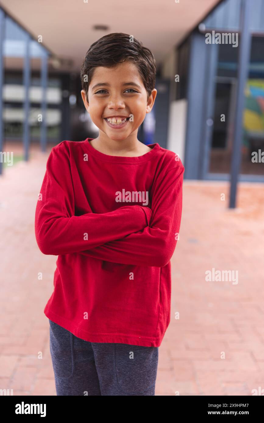 A scuola, giovane studente birazziale maschile in piedi con le braccia incrociate, sorridendo all'aperto Foto Stock