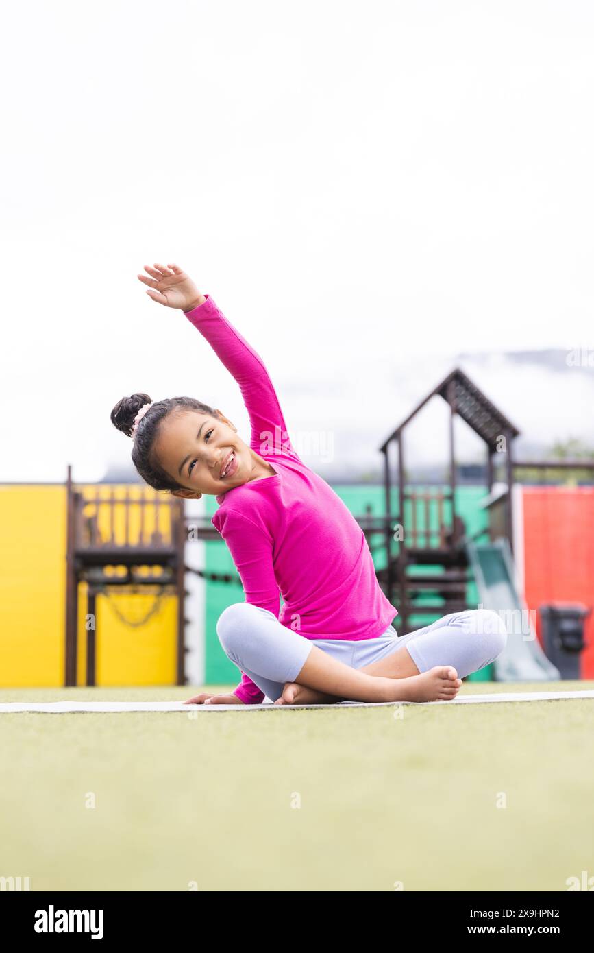 A scuola, la giovane ragazza birazziale rosa e grigia si estende sul parco giochi facendo yoga Foto Stock