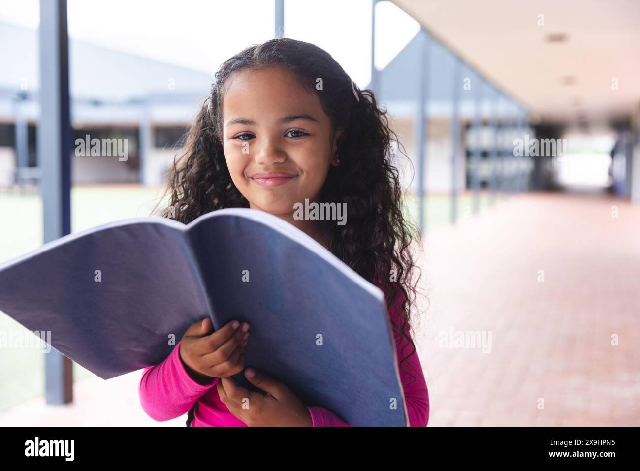 A scuola, giovane studentessa birazziale che tiene un libro, in piedi all'aperto Foto Stock