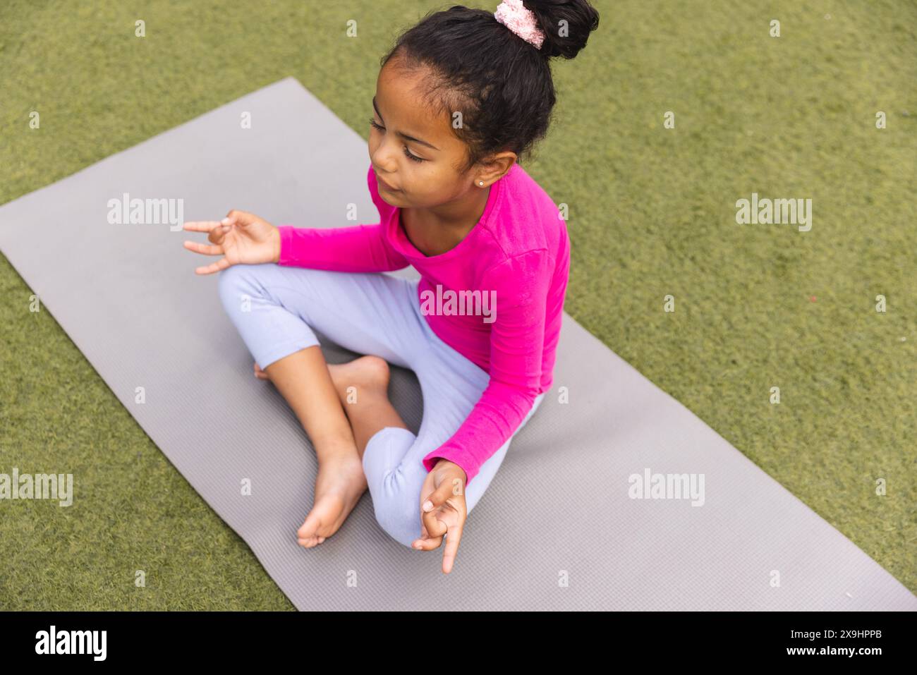 Una giovane ragazza birazziale che indossa il rosa è seduta su un tappetino da yoga all'aperto Foto Stock