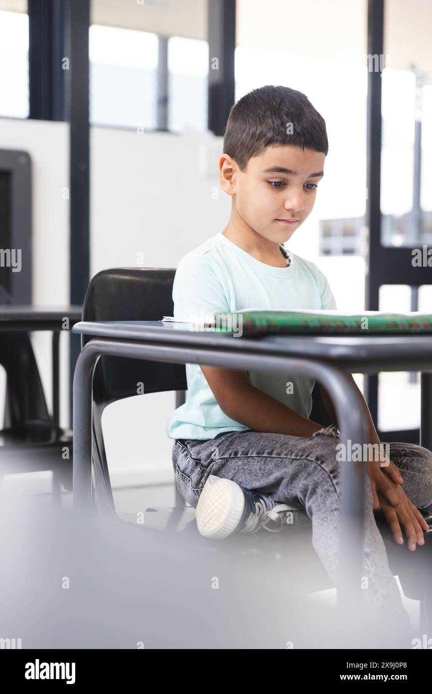 A scuola, giovane studente maschio birazziale seduto a una scrivania in classe, guardando un tablet Foto Stock