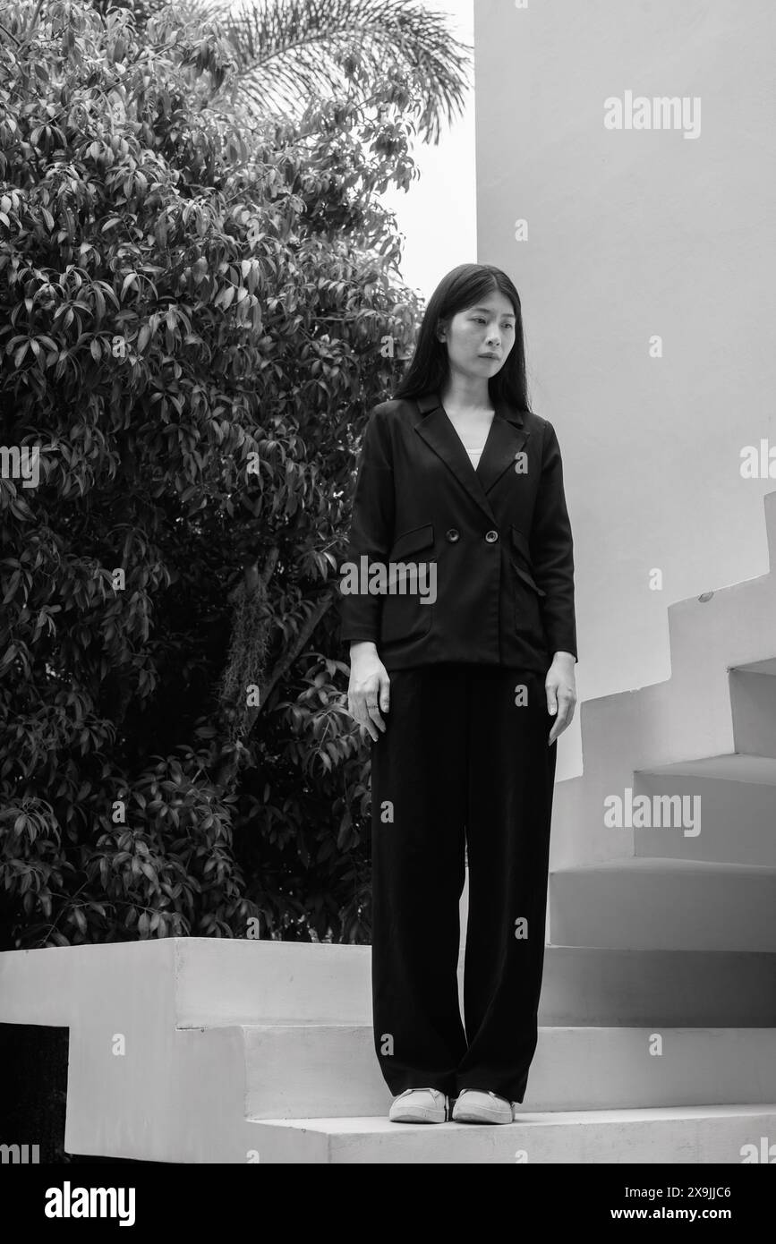 Una donna professionale in abbigliamento da lavoro si erge con sicurezza all'aperto su una scala, incorniciata da alberi sullo sfondo Foto Stock