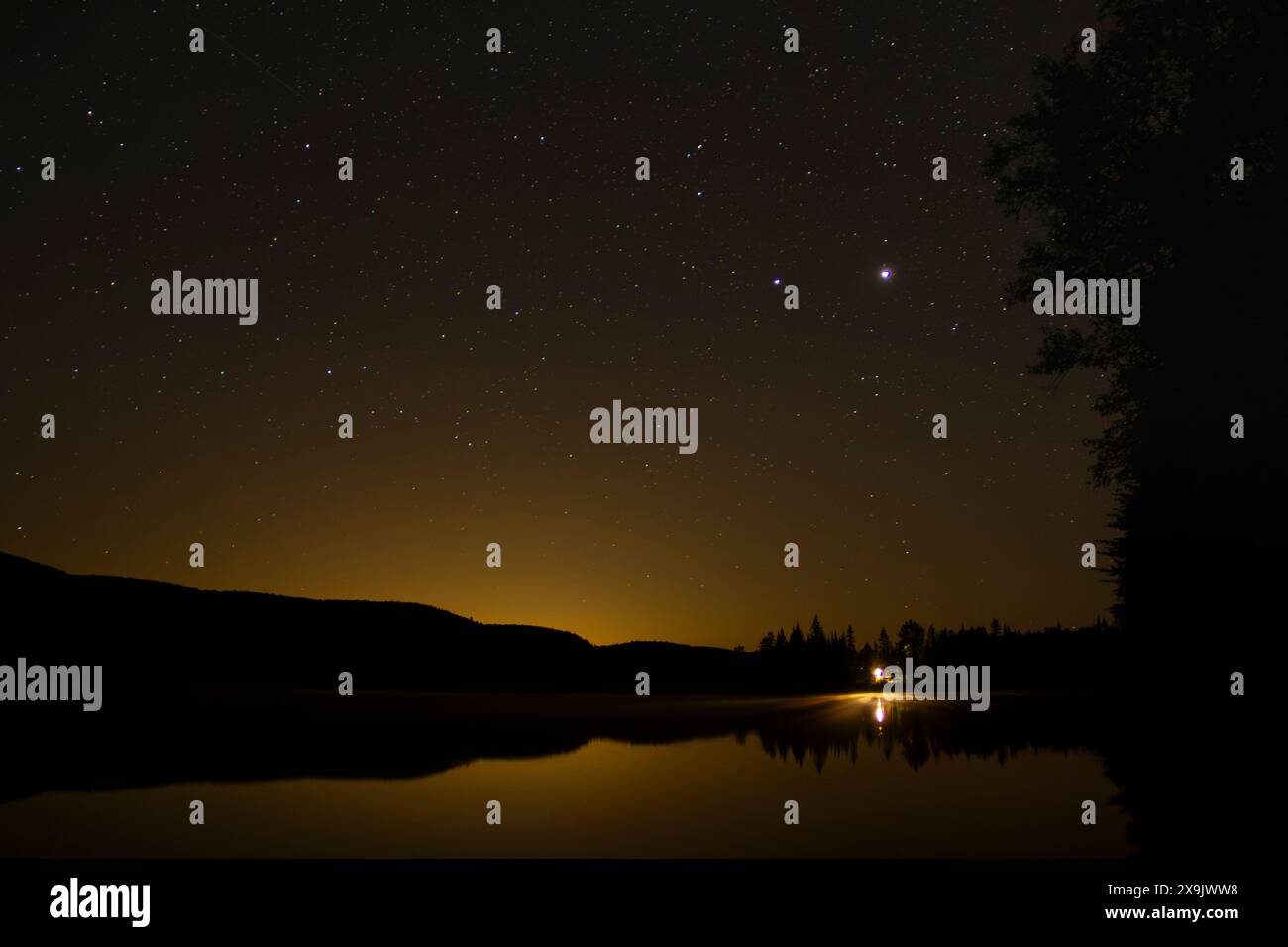 La luce di una cabina solitaria si riflette su un tranquillo lago sotto un cielo stellato nei boschi del nord del Quebec, Canada. Foto Stock
