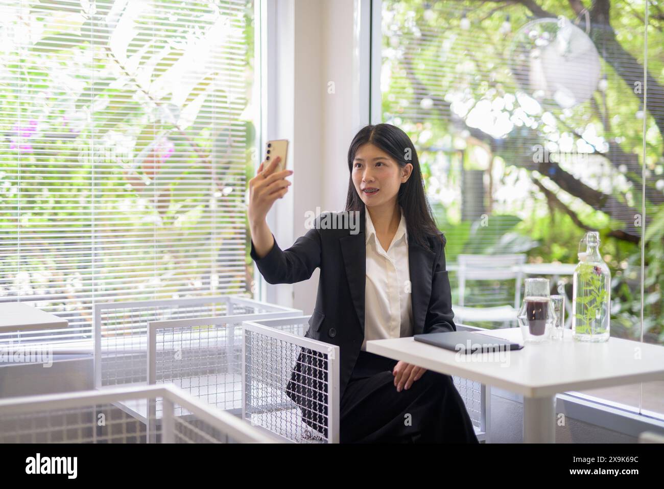 Donna d'affari asiatica professionista che lavora sul suo smartphone in un bar luminoso e moderno con caffè e vegetazione Foto Stock