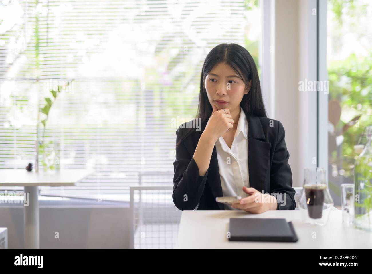 Donna d'affari asiatica professionista che lavora sul suo smartphone in un bar luminoso e moderno con caffè e vegetazione che sembrano stressati Foto Stock