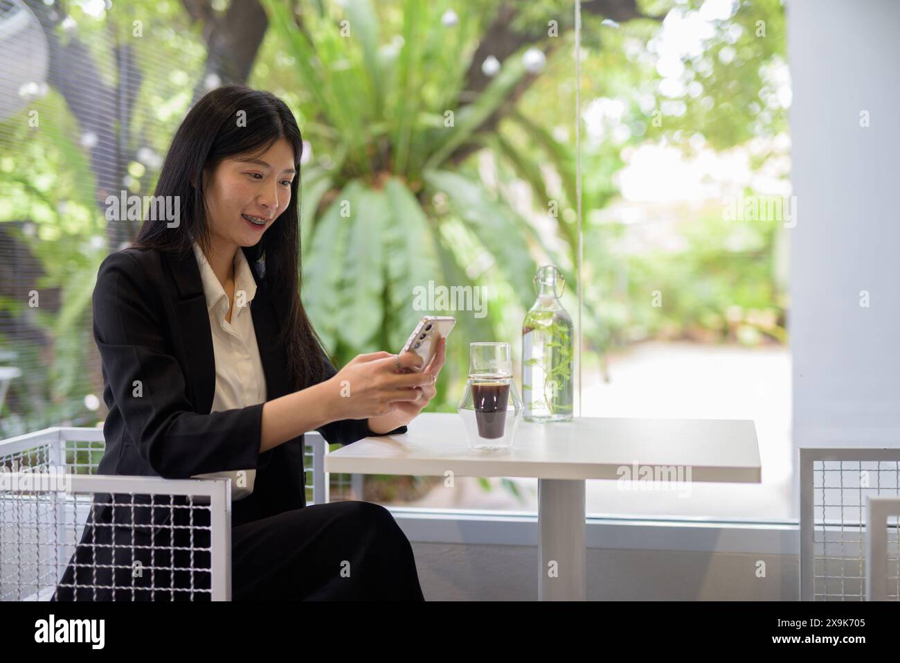 Donna d'affari asiatica professionista che lavora sul suo smartphone in un bar luminoso e moderno con caffè e vegetazione Foto Stock