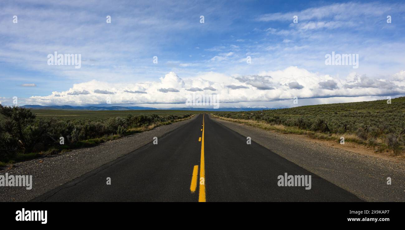 Strada aperta di asfalto balck con linea gialla nell'Oregon orientale che conduce a un ampio paesaggio con fila di nuvole bianche Foto Stock