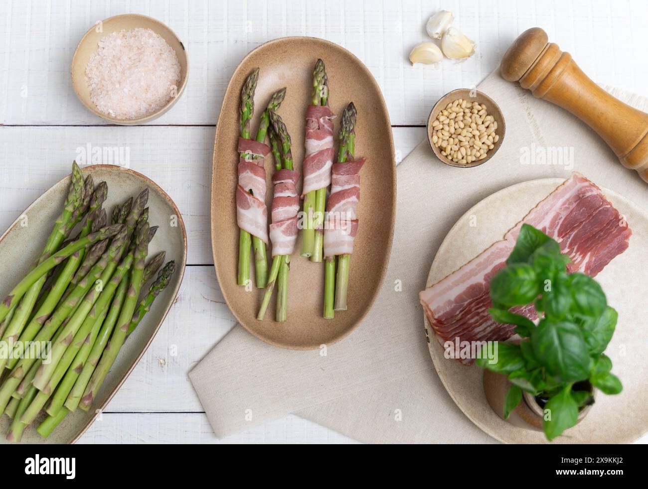 Asparagi verdi croccanti mostrati su piastre beige, pronti per il forno. Incorniciato da piatti con pancetta e altri ingredienti come aglio e pinoli Foto Stock