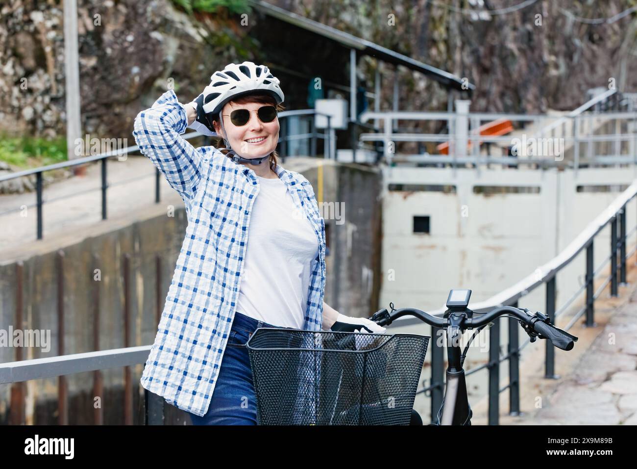 Donna matura sorridente con casco e occhiali da sole in bicicletta che si trova sul canale d'ingresso. Concetto di stile di vita attivo per persone adulte. Foto Stock