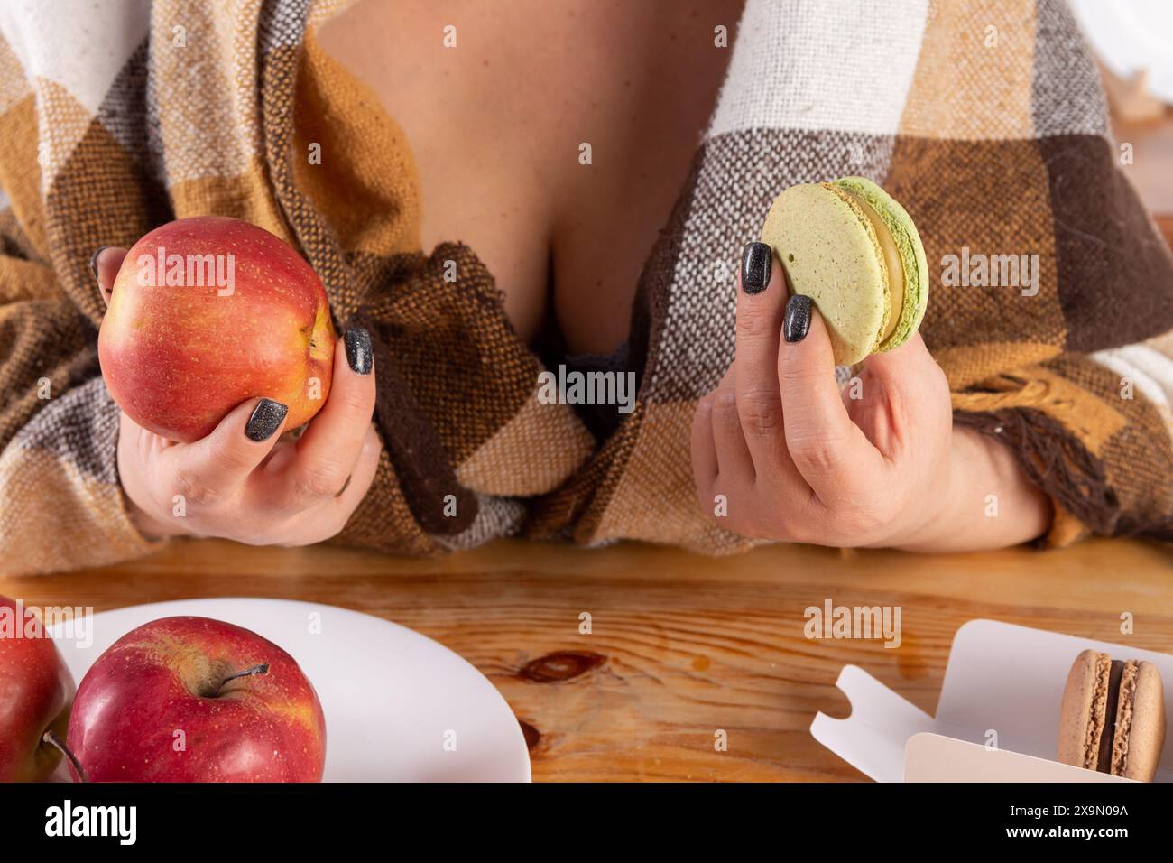 Una donna con lunghi capelli scuri indossa un cappotto marrone a quadri. Tiene una mela rossa in una mano e un biscotto verde nell'altra mano. La donna lo è Foto Stock