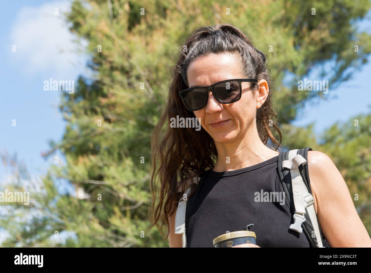 Donna in abito nero e occhiali da sole, che tiene una tazza di caffè, che gode di una giornata di sole circondati dalla natura. Foto Stock