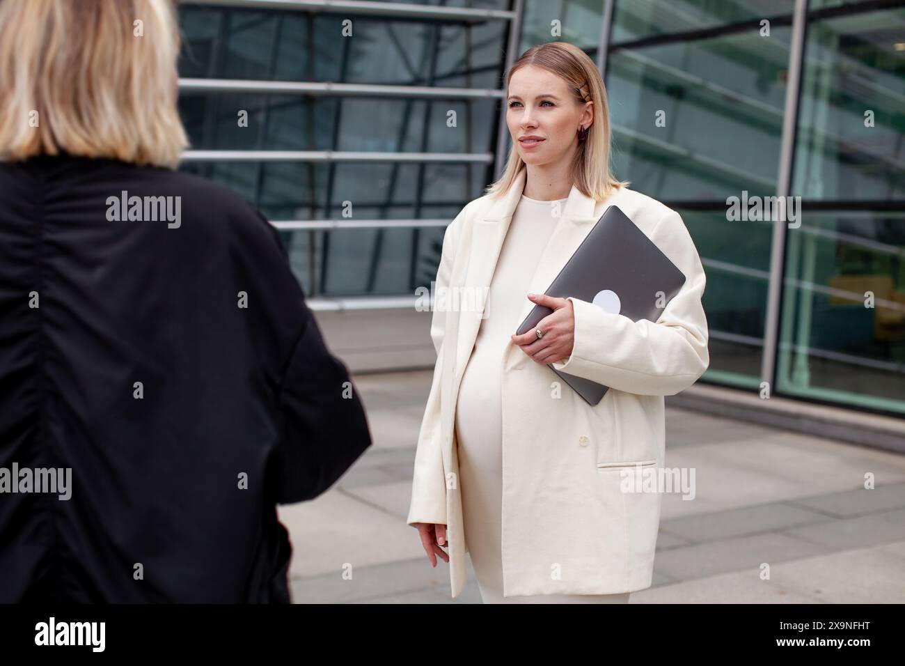 Donna d'affari incinta che parla con un collega all'aperto, tiene un laptop e indossa un elegante abbigliamento bianco per la maternità, mostrando un professionista Foto Stock