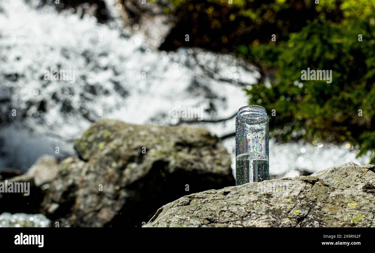 Bottiglia d'acqua riutilizzabile con acqua pulita su una pietra contro il fiume di montagna . concetto di acqua minerale naturale pulita Foto Stock