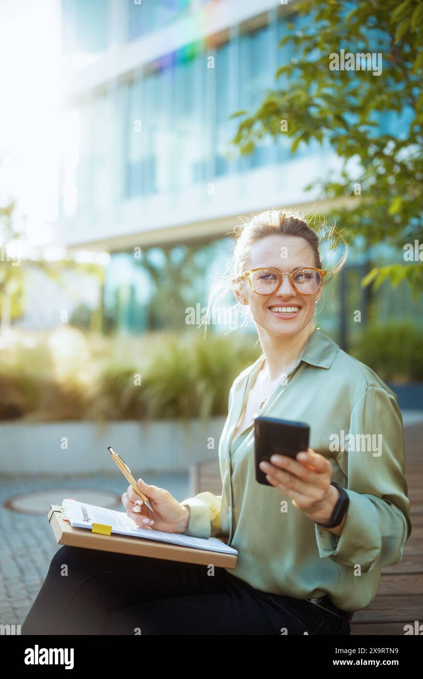 donna sorridente di 40 anni lavoratrice nel quartiere degli affari in camicetta verde e occhiali da vista con smartphone, documenti e cartella. Foto Stock