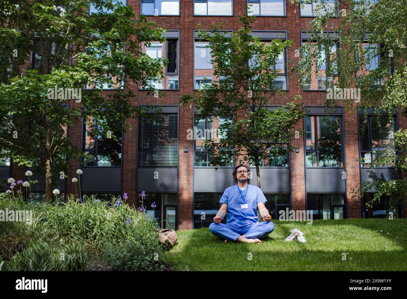 Medico seduto sull'erba, meditando e rilassandosi, dopo una giornata di lavoro intensa in ospedale. Salute mentale e equilibrio della vita per un operatore sanitario. Foto Stock