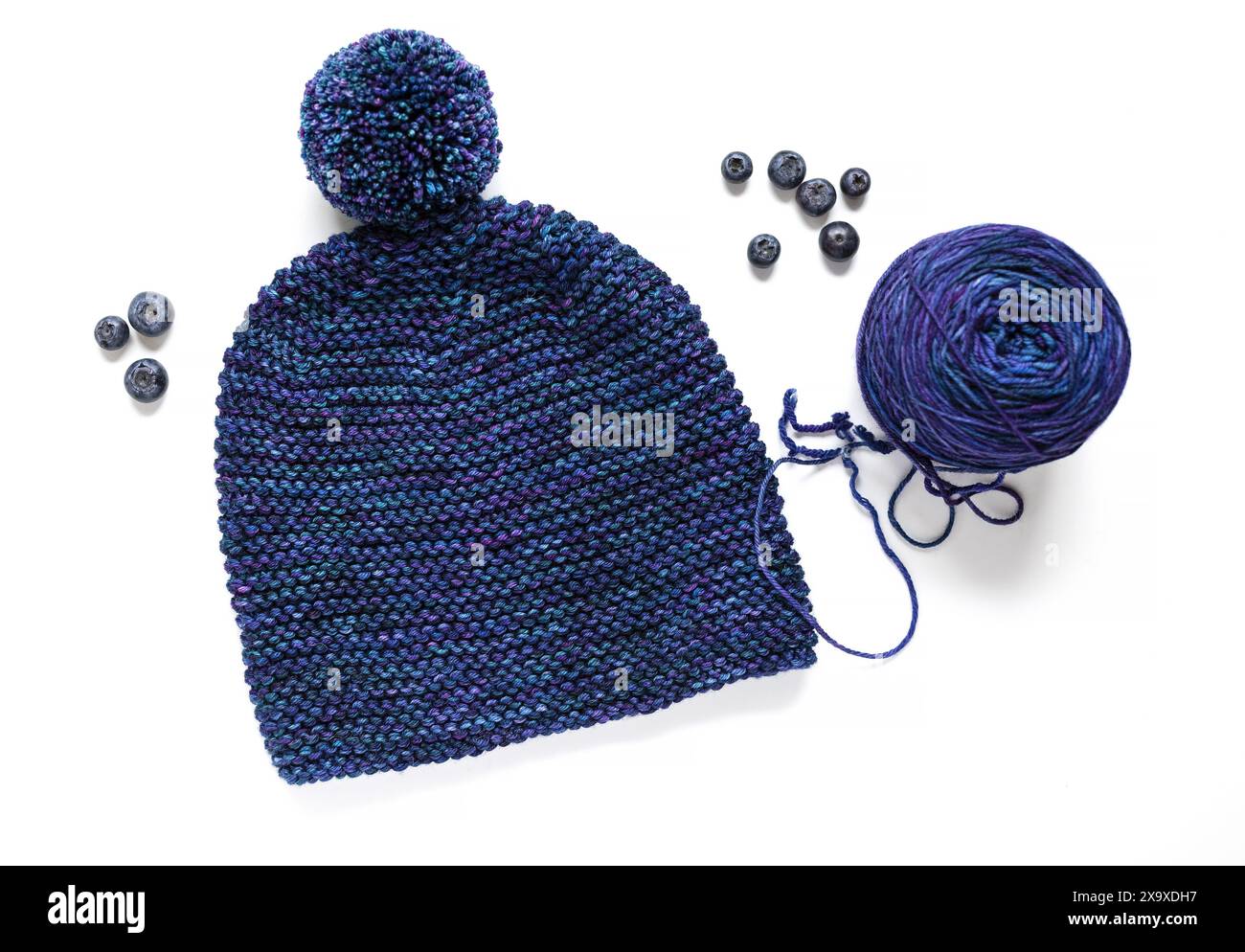 Fotografia piatta di un cappello in maglia blu con pompon e palla in filato di lana tinta a mano su sfondo bianco Foto Stock