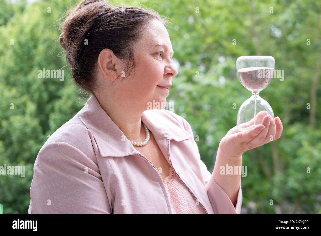 Donna matura, 55 anni, vestita di rosa, con clessidra, abbracciando il tempo di bellezza, riflessione sulla vita, grazia invecchiamento e importanza assaporare i momenti della vita, Cheris Foto Stock