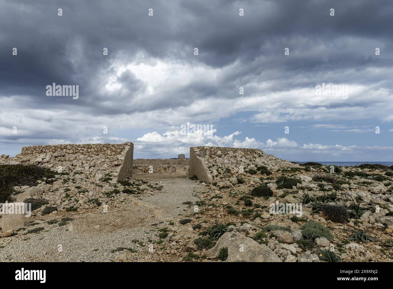 Una batteria antiaerea abbandonata a Cabo de Cavalleria con nuvole scure e drammatiche in alto. Le rovine storiche sorgono in un paesaggio aspro. Foto Stock