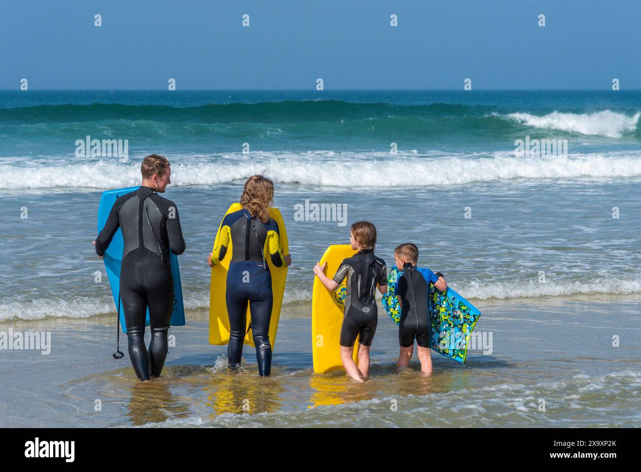 Una giovane famiglia di vacanzieri che si gode il clima caldo trasportando le proprie tavole da bodyboard nel mare alla spiaggia Fistral di Newquay. Foto Stock