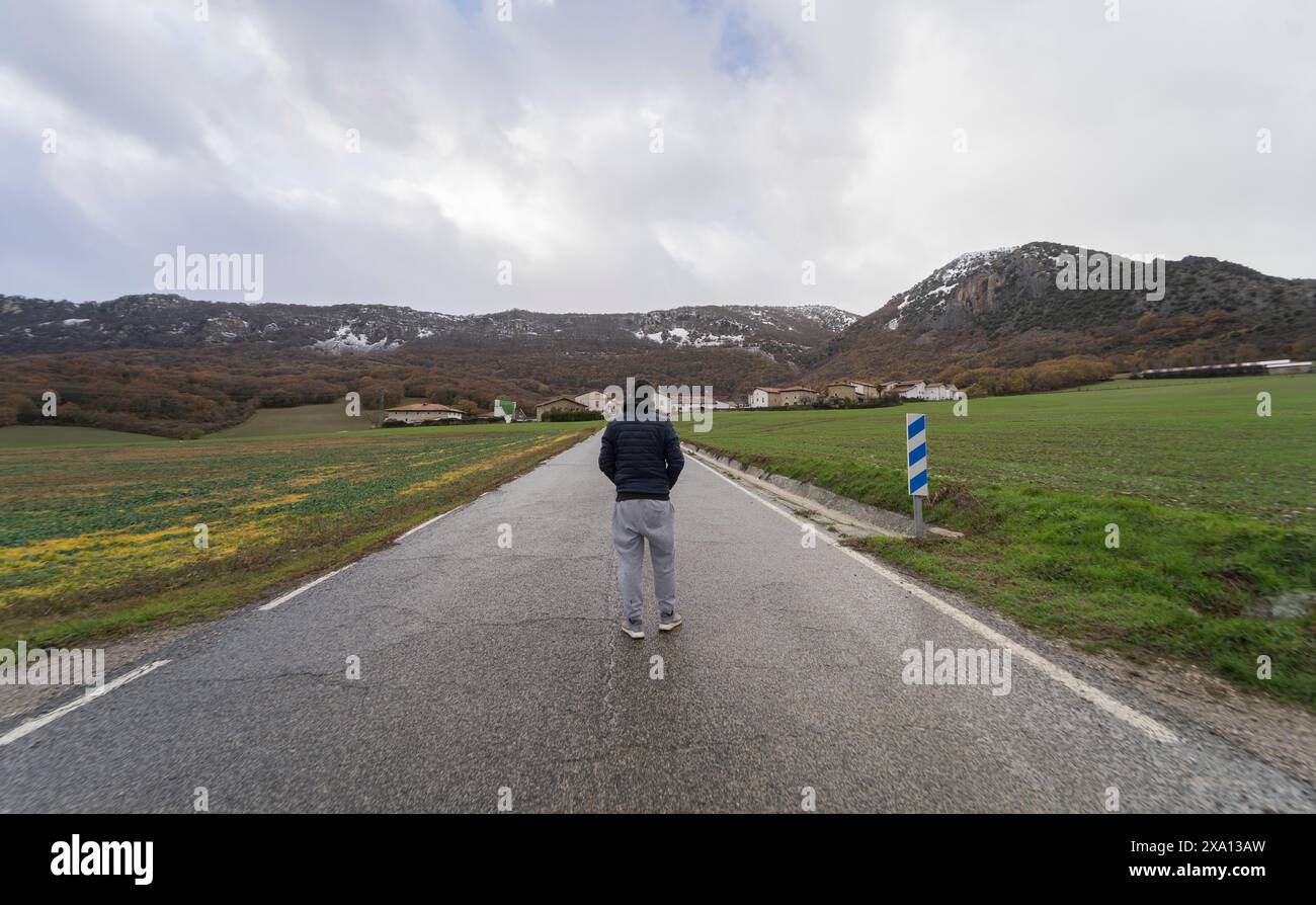 Un uomo cammina lungo una strada umida che conduce a un villaggio di Navarra, in Spagna, mentre guarda le montagne innevate e il cielo coperto. Valle Baztan Foto Stock