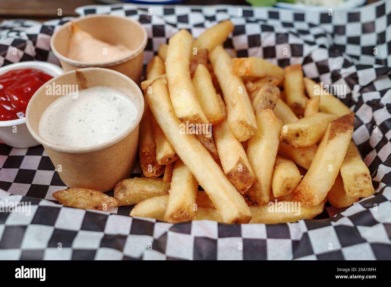 Uno shot ravvicinato di una porzione di patatine fritte dorate e croccanti presentate su una carta a scacchi. Le patatine sono accompagnate da tre salse. Foto Stock