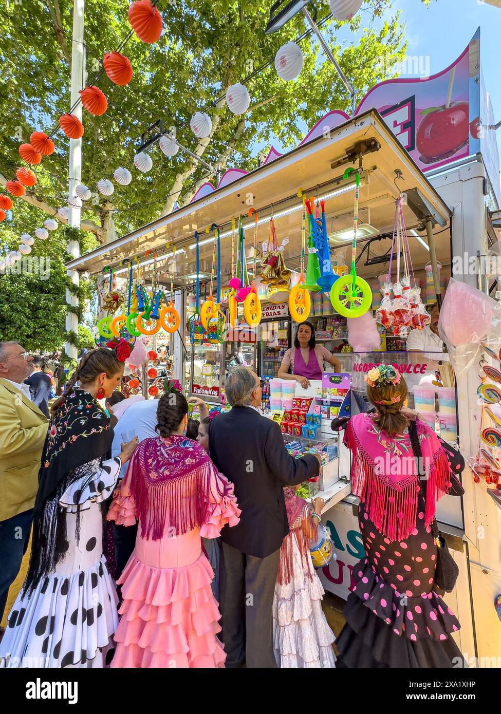 I visitatori del festival indossano abiti tradizionali al Feira de Abril di Siviglia, facendo la fila a un venditore di dolci durante il festival della durata di una settimana Foto Stock