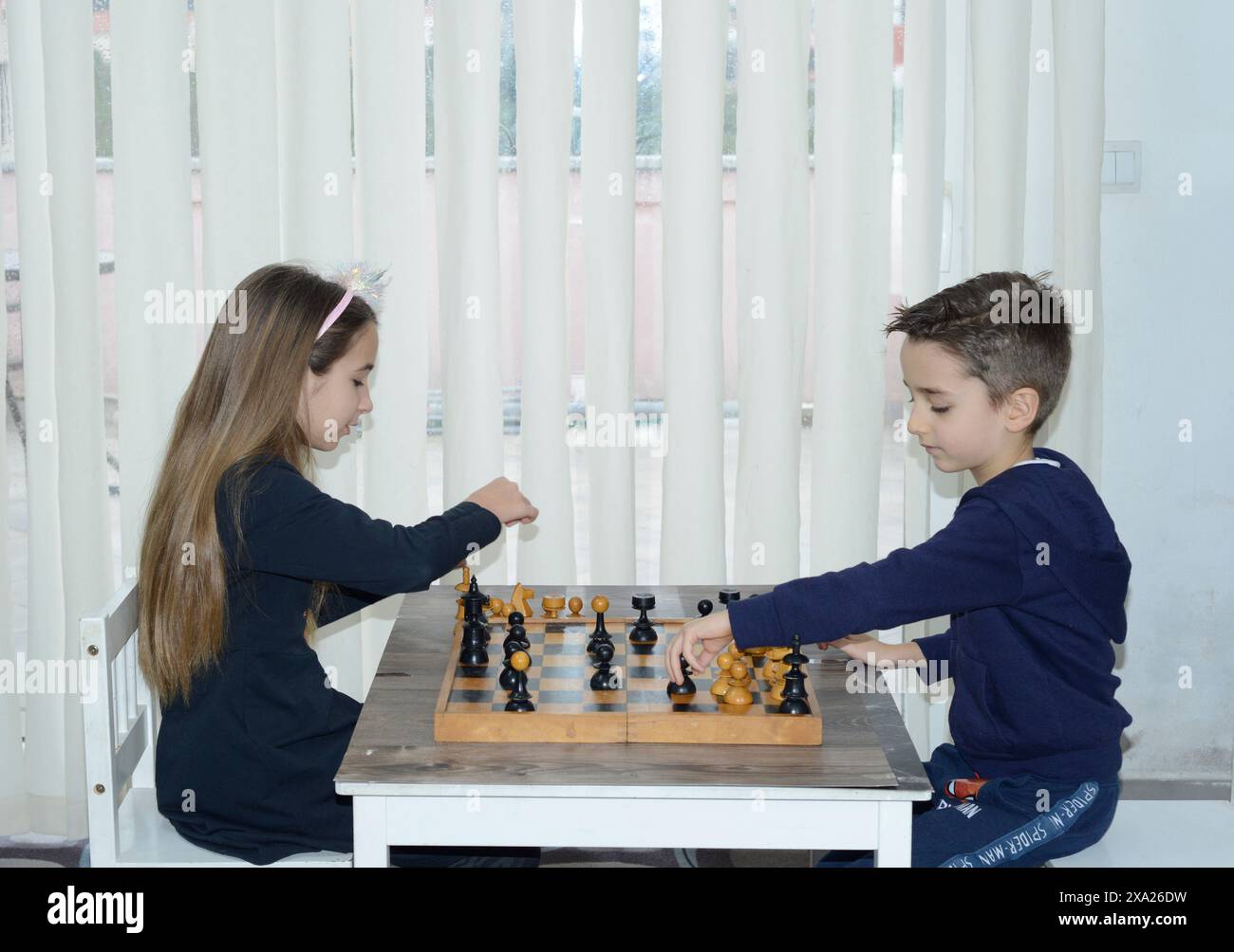 Due bambini che giocano a scacchi a un tavolo con una tenda sullo sfondo Foto Stock