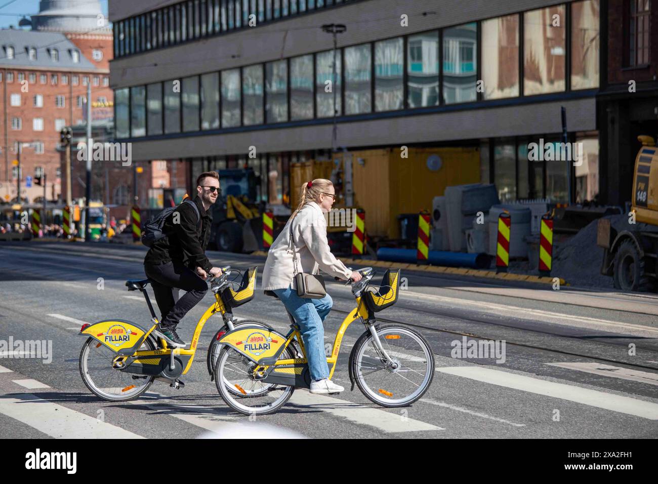 Coppia che attraversa Siltasaarenkatu in sella a HSL City Bikes o Alepa Bikes nel quartiere Hakaniemi di Helsinki, Finlandia Foto Stock