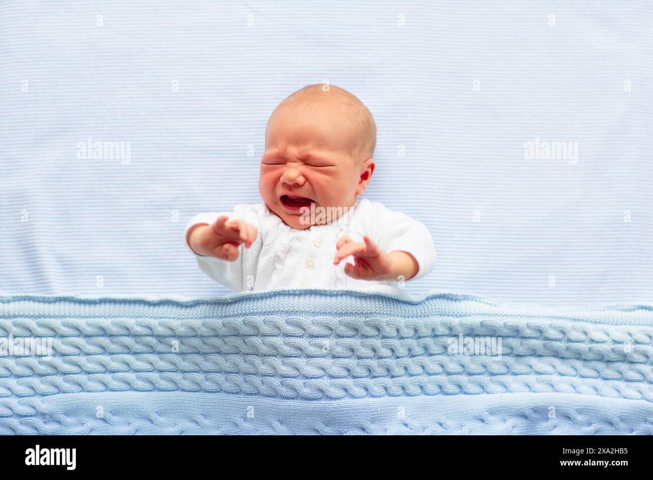 Bambino appena nato in lacrime a letto. Neonato che dorme sotto una coperta a maglia blu. I bambini piangono. Biancheria da letto per bambini. Sonnellino per neonati nel letto. Foto Stock