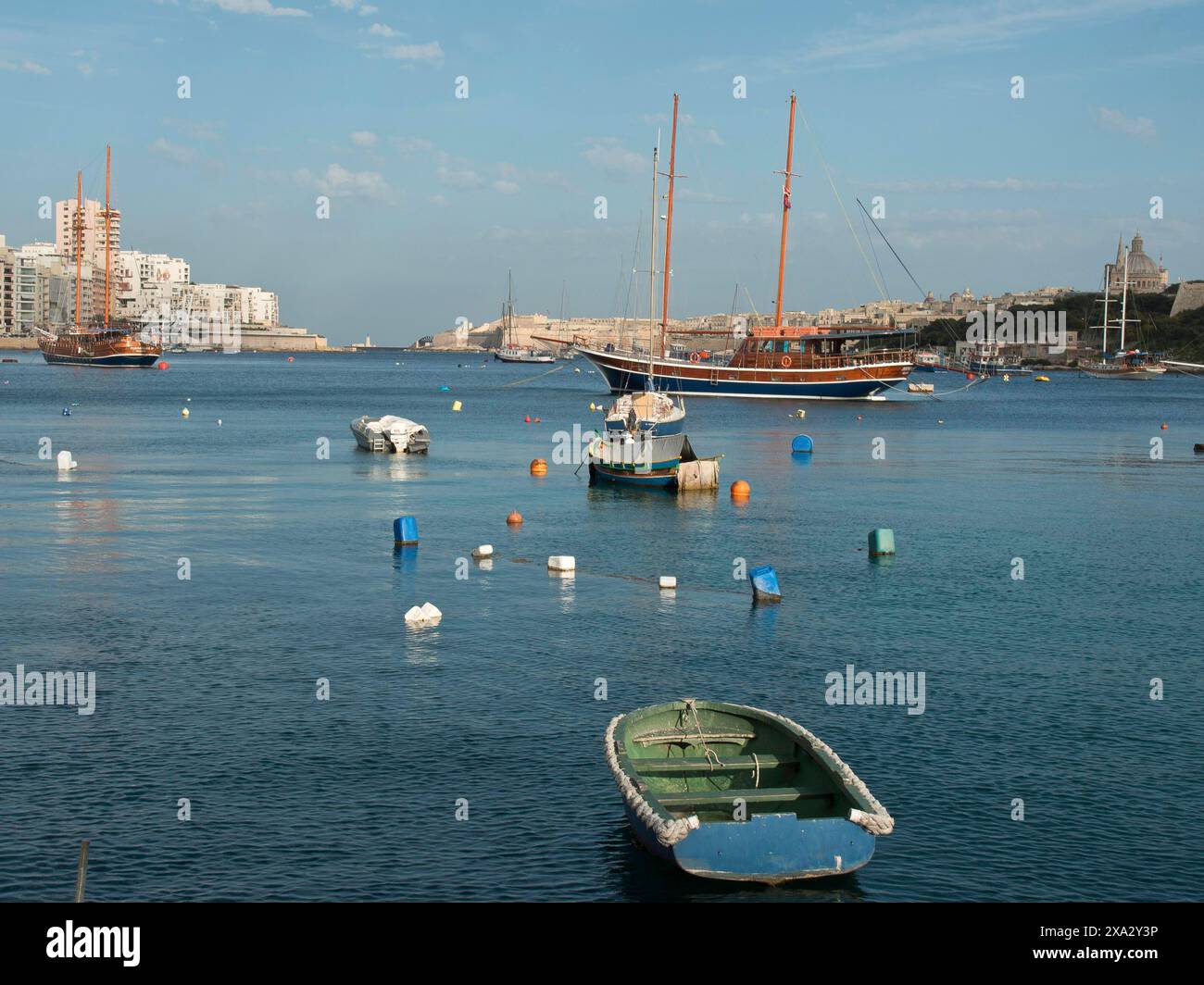 Diverse barche e una grande barca a vela sul mare calmo di fronte a una città storica e boe colorate, la Valletta, Malta Foto Stock