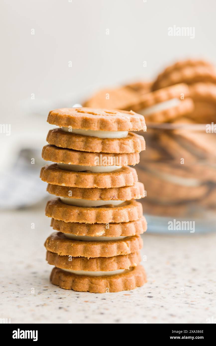 Una pila di otto biscotti ripieni di panna si trova su un ripiano, con uno sfondo sfocato di altri biscotti in una ciotola di vetro. I biscotti hanno un marrone chiaro Foto Stock