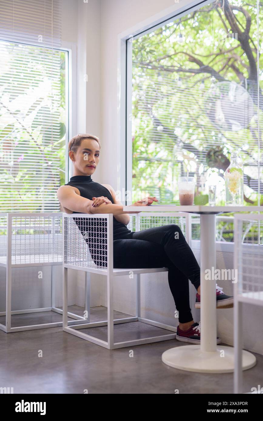 Ritratto a figura intera di un bellissimo ragazzo gay asiatico, sostenitore della comunità LGBT, con baffi che indossano rossetto e tuta in un bar luminoso e moderno con i Foto Stock