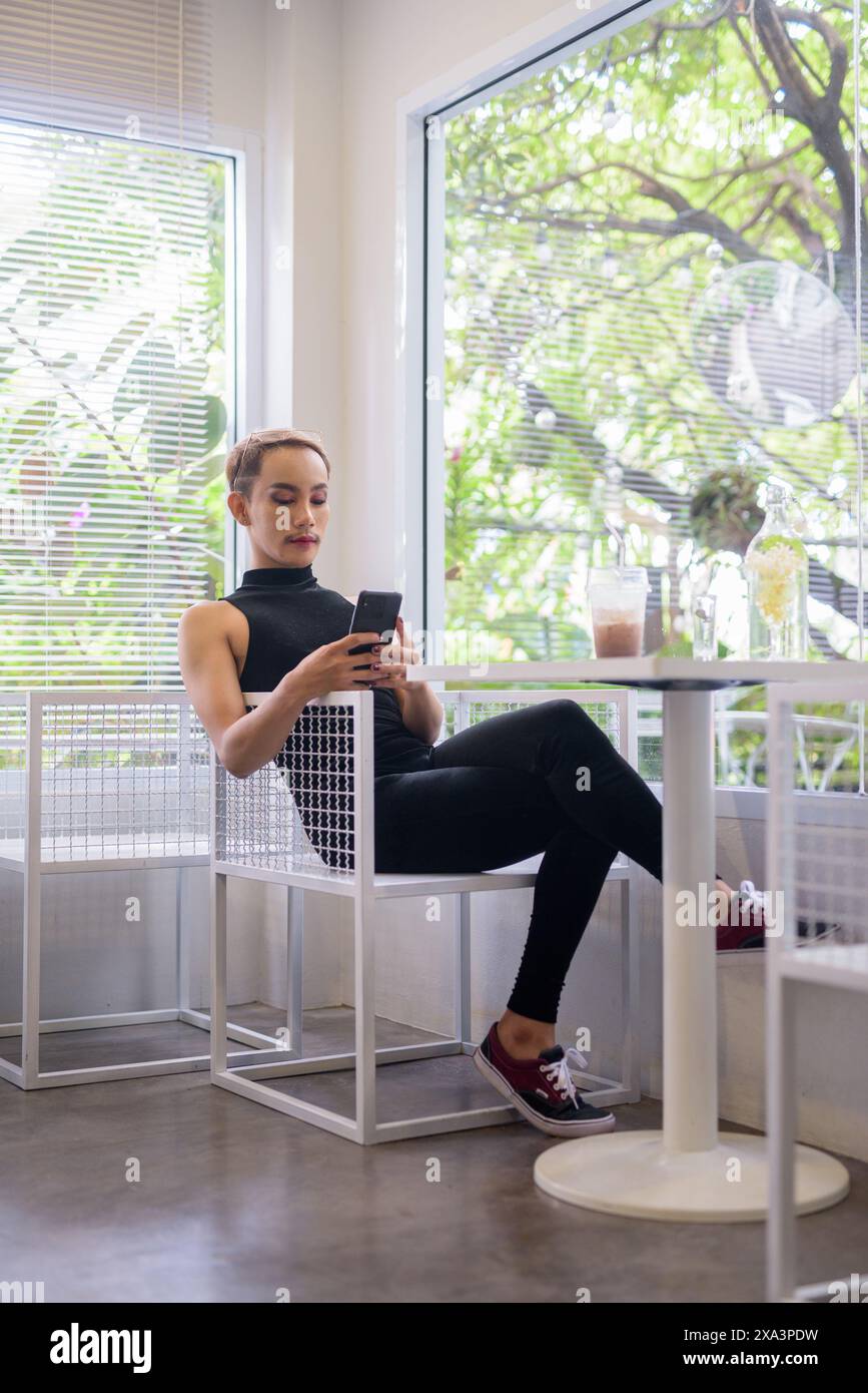 Ritratto a figura intera di un bellissimo ragazzo gay asiatico, sostenitore della comunità LGBT, con baffi che indossano rossetto e tuta in un bar luminoso e moderno con i Foto Stock