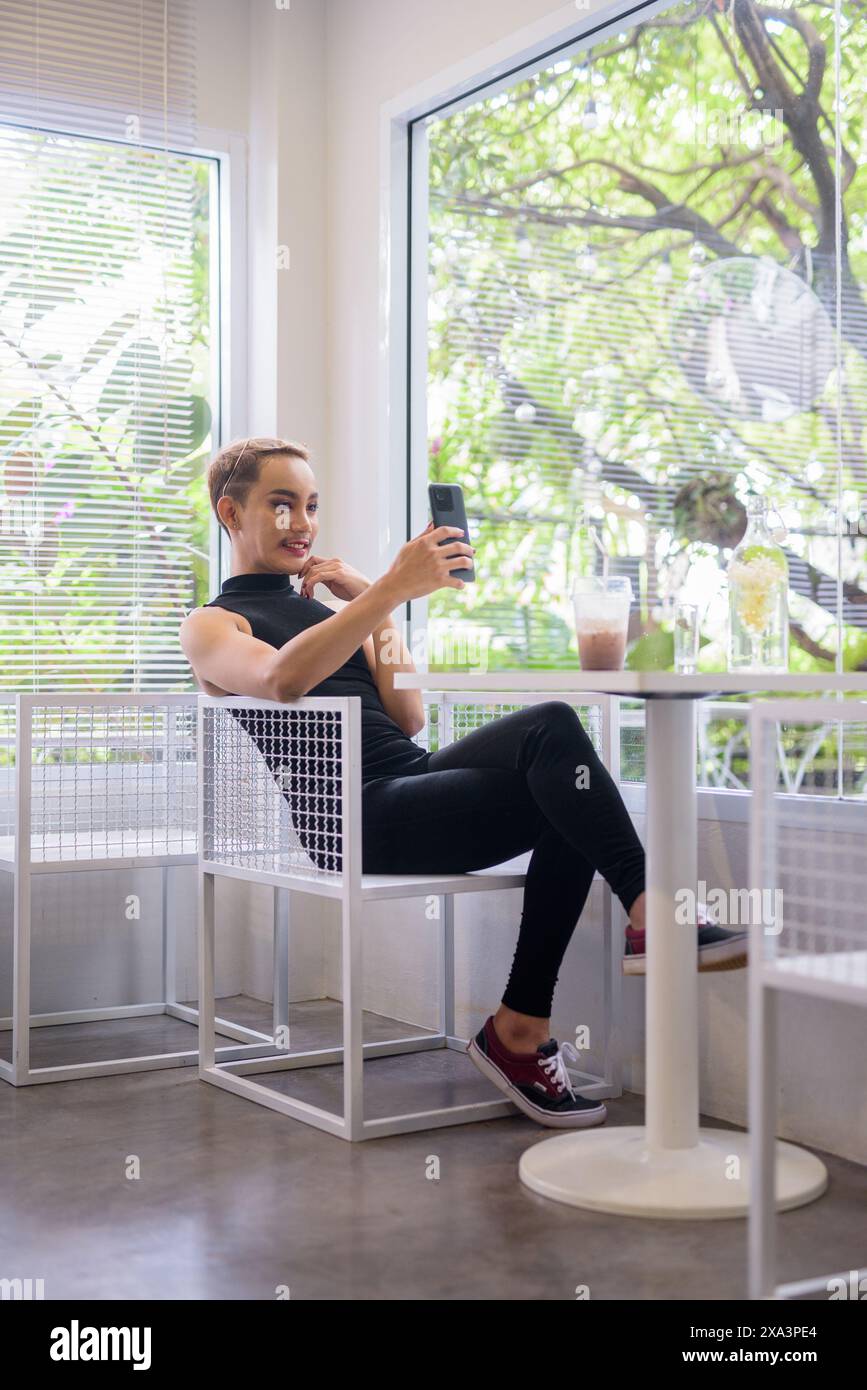 Ritratto a figura intera di un bellissimo ragazzo gay asiatico, sostenitore della comunità LGBT, con baffi che indossano rossetto e tuta in un bar luminoso e moderno con i Foto Stock