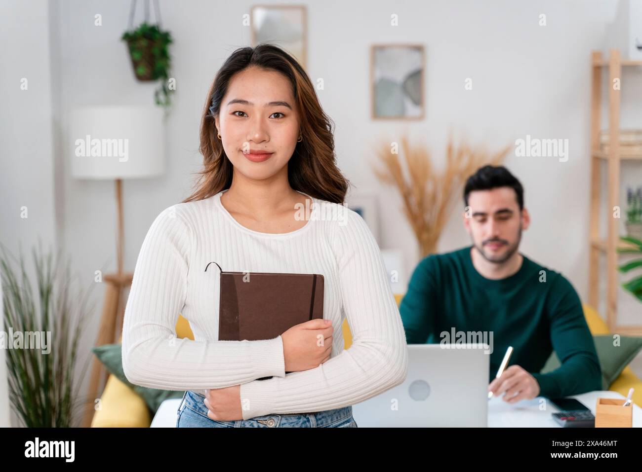 Giovane donna con un libro in piedi in un ufficio, uomo che lavora al portatile dietro di lei. Foto Stock