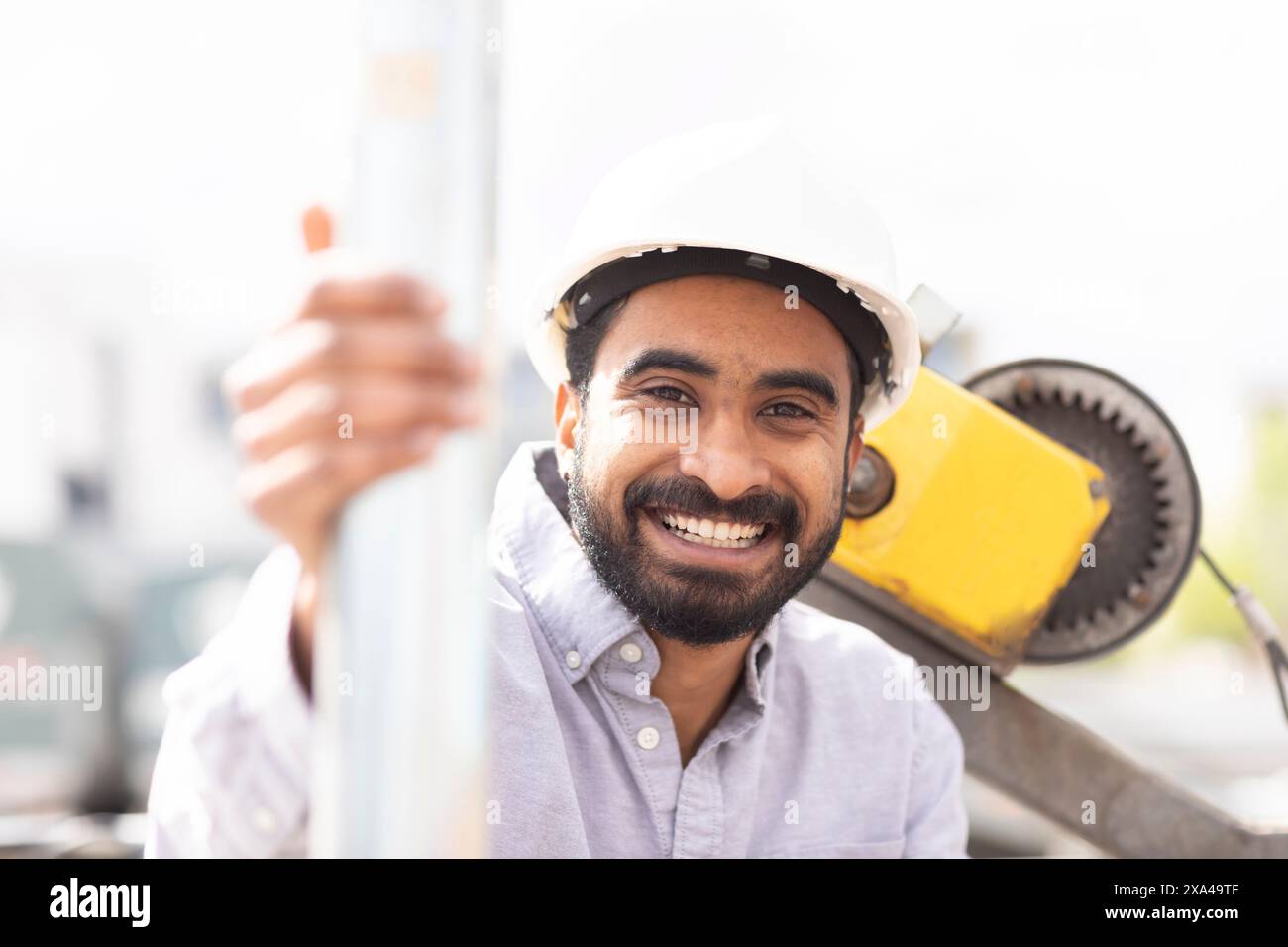 Un uomo sorridente con un elmetto bianco si appoggia su un palo metallico in un cantiere edile. Foto Stock