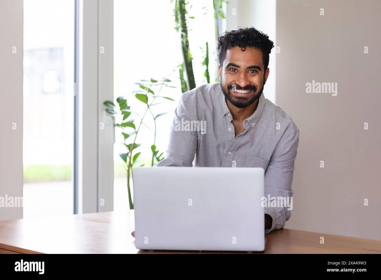 Un uomo sorridente con la barba a un tavolo di legno in una stanza ben illuminata, che lavora su un computer portatile Foto Stock