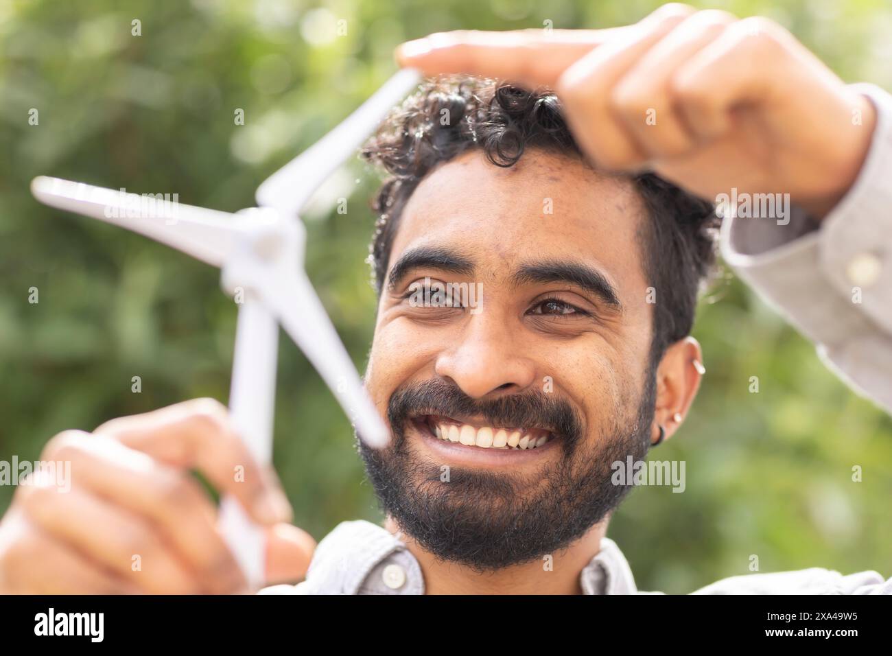 Un uomo sorridente con capelli ricci tiene un piccolo modello di turbina eolica bianca sopra la testa su uno sfondo verde sfocato. Foto Stock