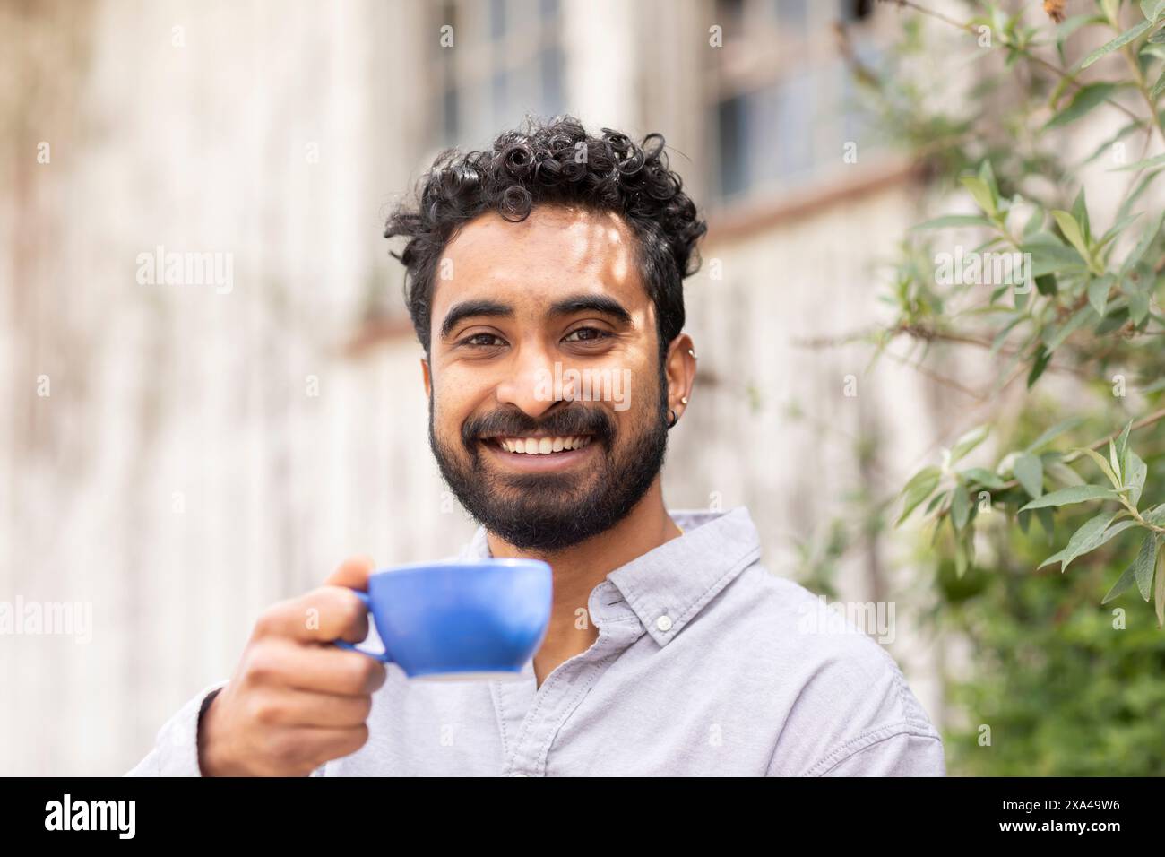 Un uomo sorridente con i capelli ricci tiene una tazza blu verso la fotocamera, davanti a uno sfondo sfocato Foto Stock
