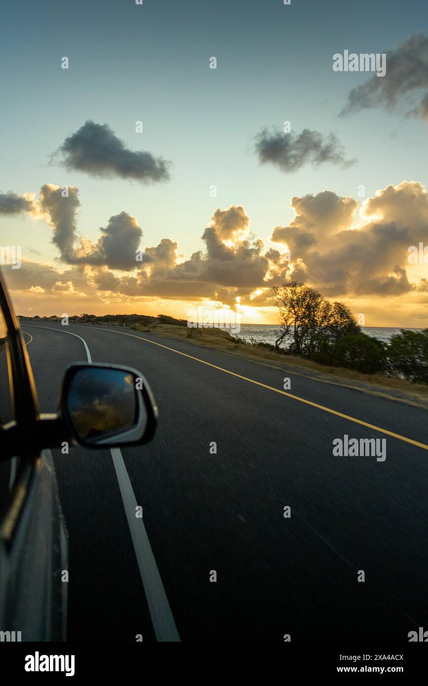 Una scena di viaggio su strada catturata dalla prospettiva di un passeggero in un veicolo in movimento, che mostra uno specchio laterale, la strada aperta davanti a te e una splendida alba o tramonto con pittoresche nuvole sullo sfondo. Foto Stock