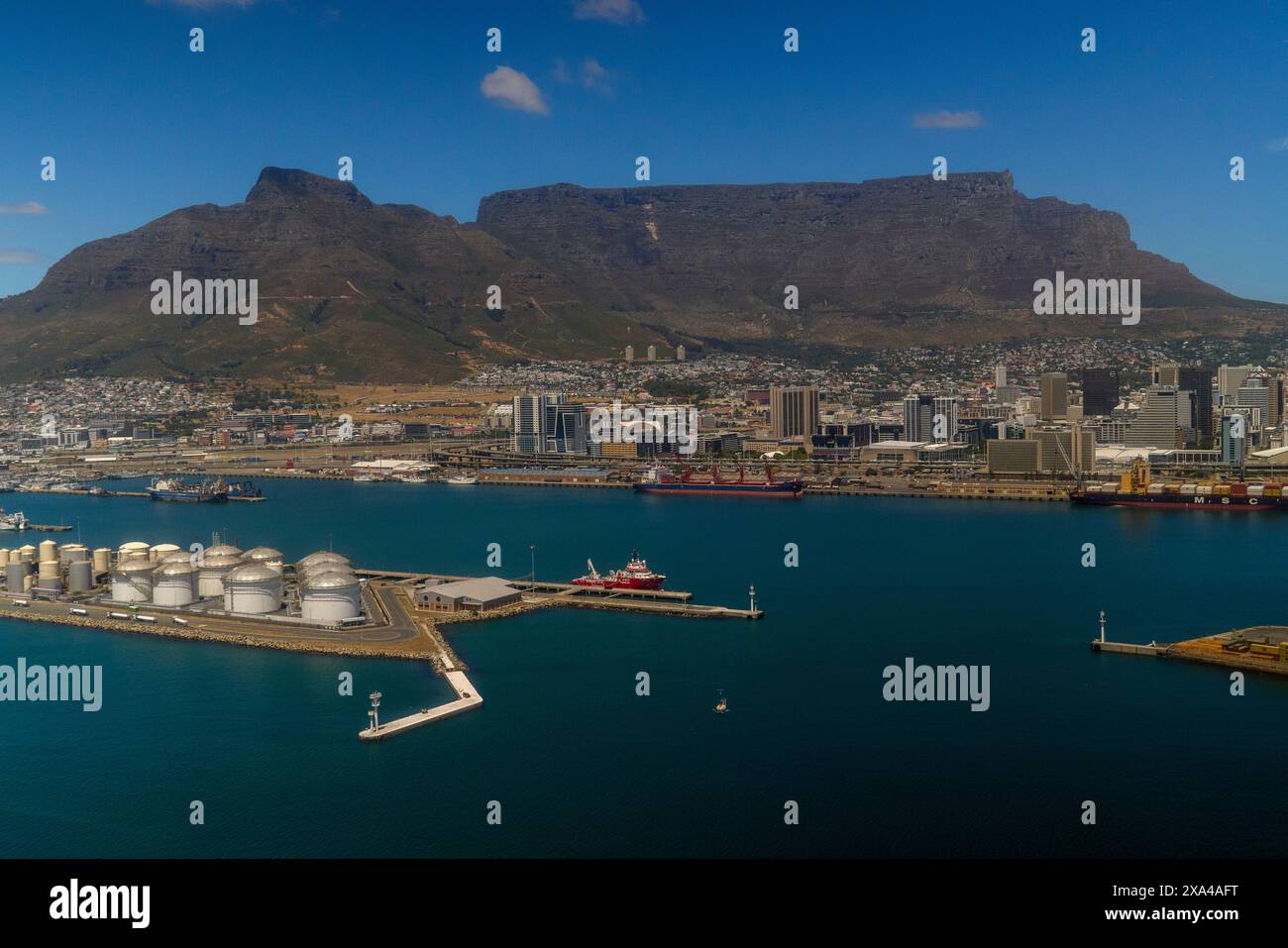 Una vista aerea di un paesaggio urbano costiero con una grande montagna sullo sfondo, un porto con navi e serbatoi di stoccaggio in primo piano e cieli azzurri chiari sopra la testa. Foto Stock