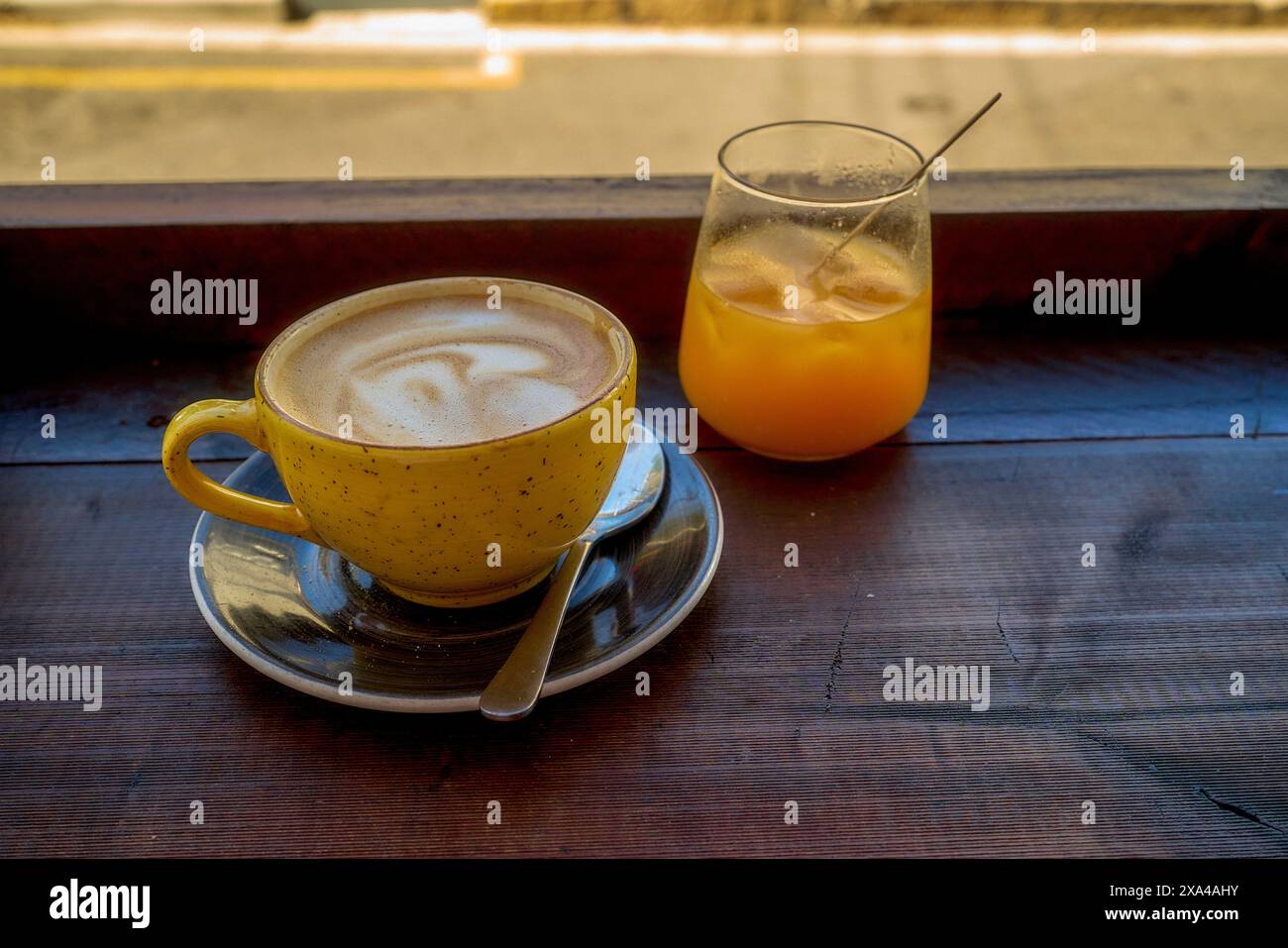 Una tazza di cappuccino con schiuma artistica accanto a un bicchiere di succo d'arancia su un tavolo di legno vicino alla finestra, che riflette un'atmosfera accogliente per la colazione. Foto Stock