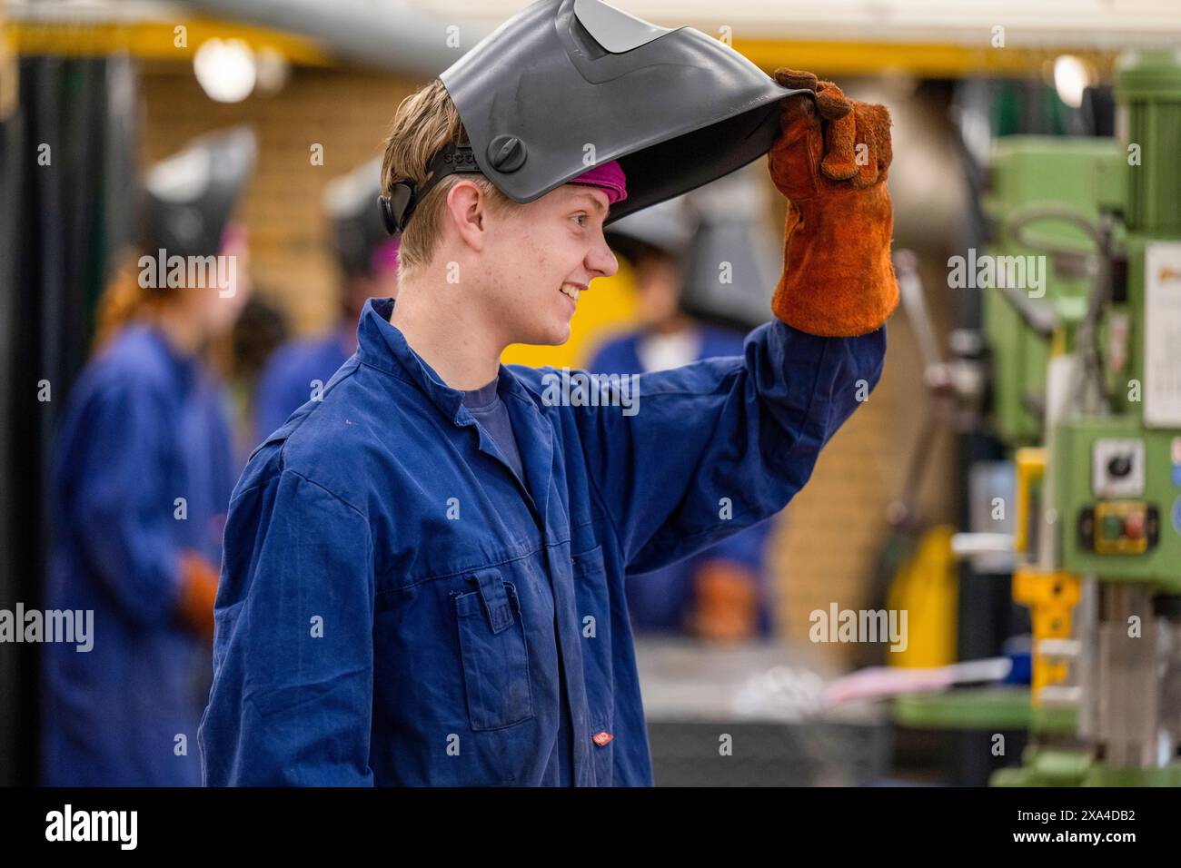Un giovane sorridente che indossa un'uniforme da lavoro blu sta sollevando la maschera di saldatura con una mano con i guanti in un ambiente industriale. Foto Stock