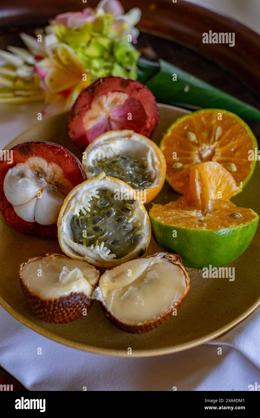 Un piatto con un assortimento di frutta tropicale, tra cui arancia affettata, frutto della passione e altri, servito su un vassoio di legno con fiori colorati sullo sfondo. Foto Stock