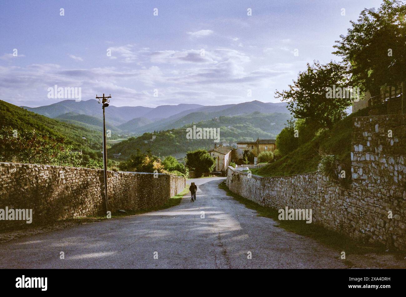 Una vista tranquilla di una vecchia strada rurale con pareti di pietra su entrambi i lati che conducono verso colline verdi ondulate, con una sola persona che cammina in lontananza sotto un cielo morbido. Foto Stock