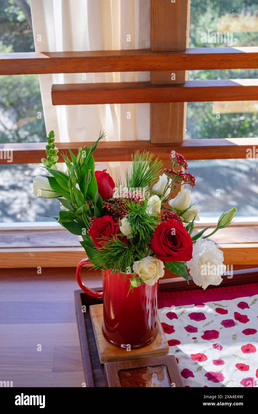 Un vivace bouquet di fiori con rose rosse e tulipani bianchi in una tazza rossa, collocata su una superficie di legno vicino ad una finestra con tende a vista e una vista del verde esterno. Foto Stock