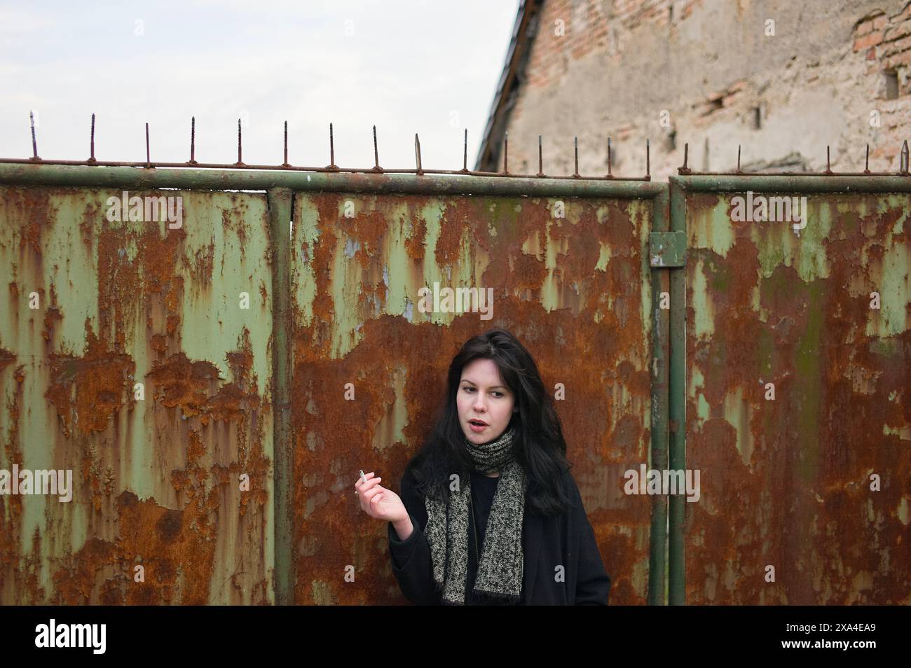Una donna si trova di fronte a un cancello di metallo arrugginito con punte sulla parte superiore, con un muro di mattoni sullo sfondo, che crea una sorprendente espressione facciale. Foto Stock