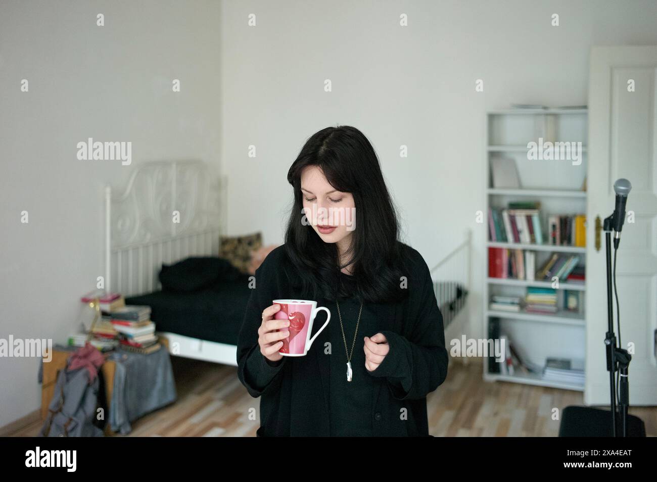 Una donna sta in una camera da letto con una tazza, con una libreria, un letto e attrezzature musicali sullo sfondo. Foto Stock