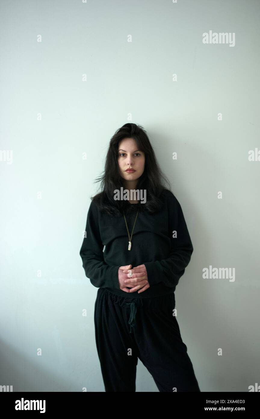 Una donna si staglia contro una parete di colore chiaro indossando abiti scuri e casual, guardando la fotocamera con un'espressione neutra. Foto Stock